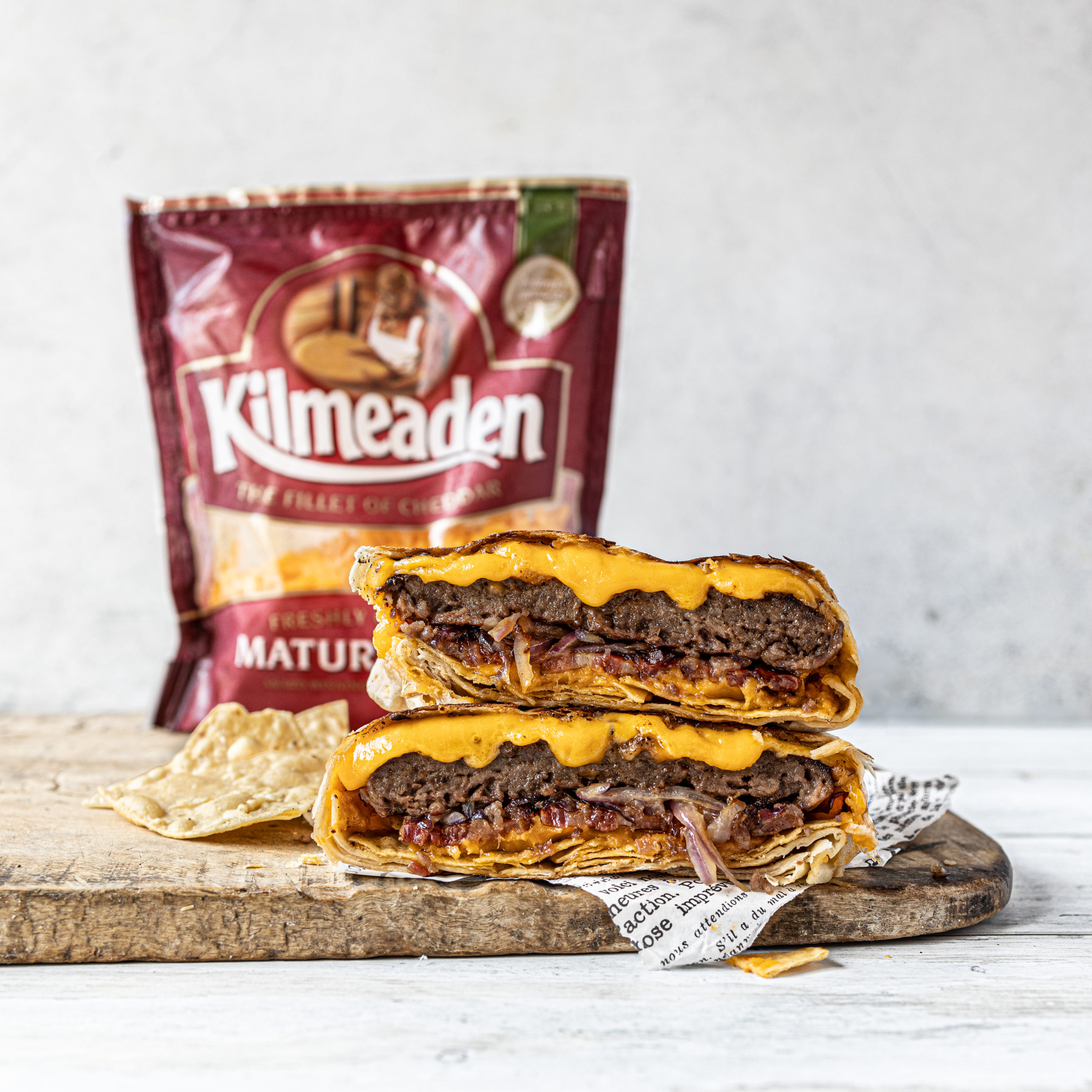 Toasted wrap with burger, bacon and mature red Kilmeaden cheddar cheese on wooden chopping board with pouch of red Kimeaden mature grated cheese in the background.