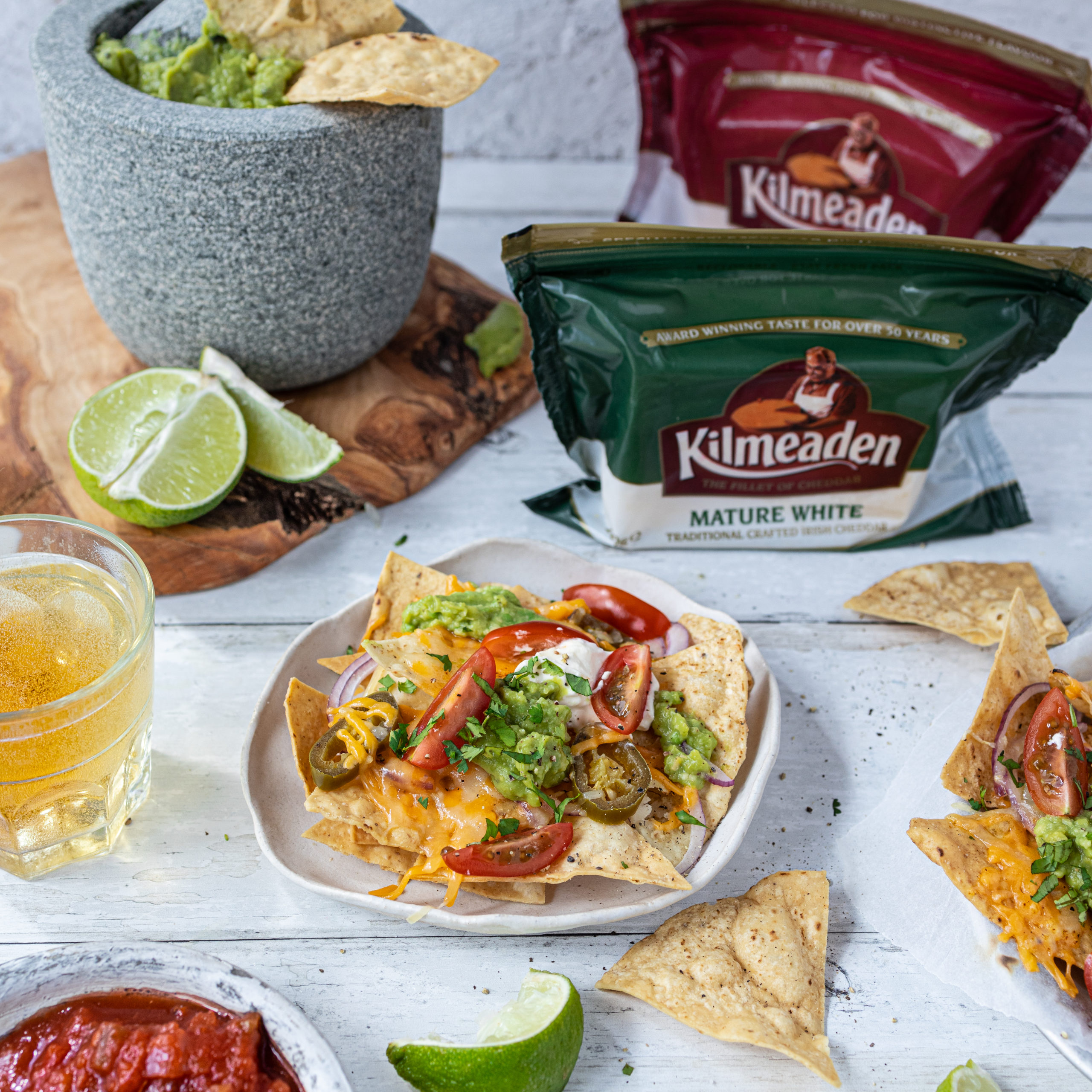 Nachos with melted grated mature red Kilmeaden cheee, lettuce, peppers, jalapenos, sour cream, guacamole, onion and lime in the centreground with ingredients scattered around the table it is on. Kilmeaden mature white and mature red cheese block in the background.