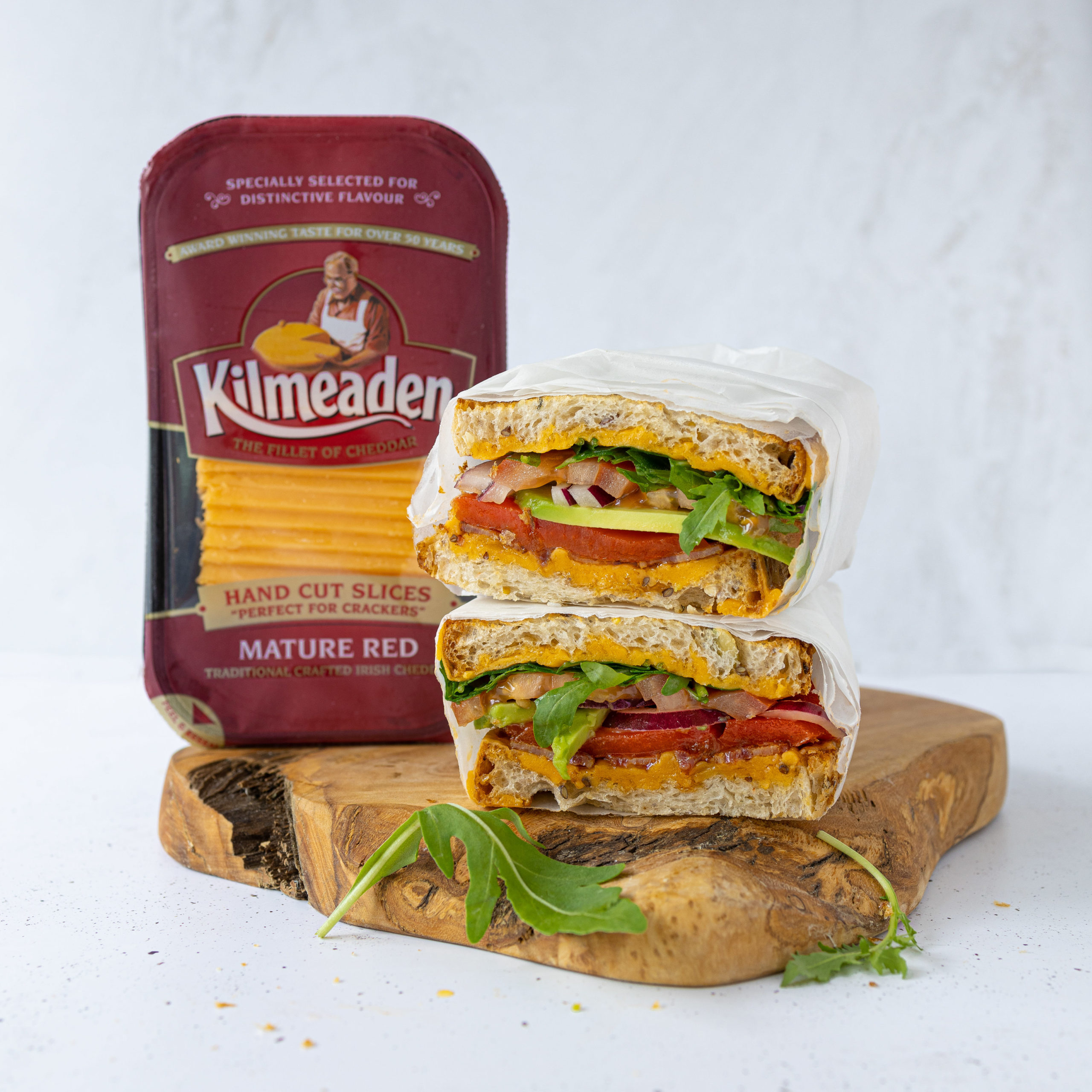 Toasted sandwich with lettuce, onion, tomato, and kilmeaden red cheddar cheese on wooden chopping board beside pack of Kilmeaden Mature Red Hand Cut Cheese slices.