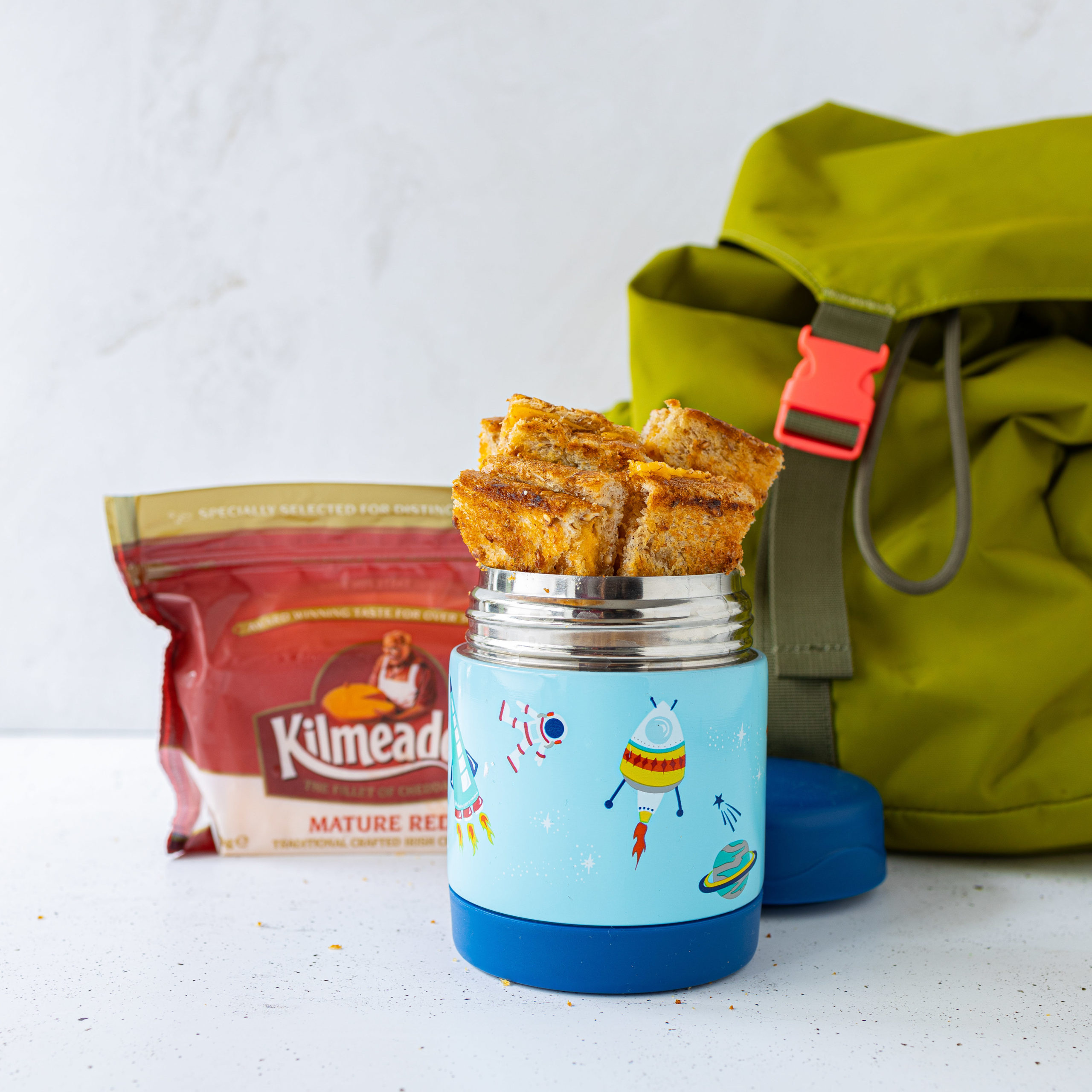Toasted Cheddar Cheese sandwich in soup container with Kilmeadan Mature red cheese block in the background along side green backpack.